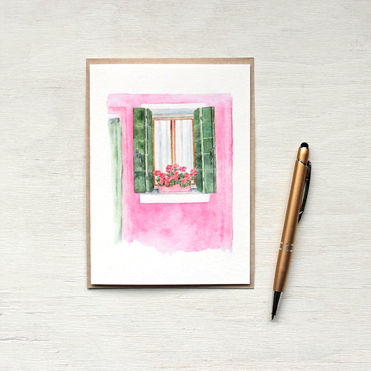 Note card featuring a pink house with green shutters and matching geraniums on the windowsill. A scene from Burano, Italy. Artist Kathleen Maunder