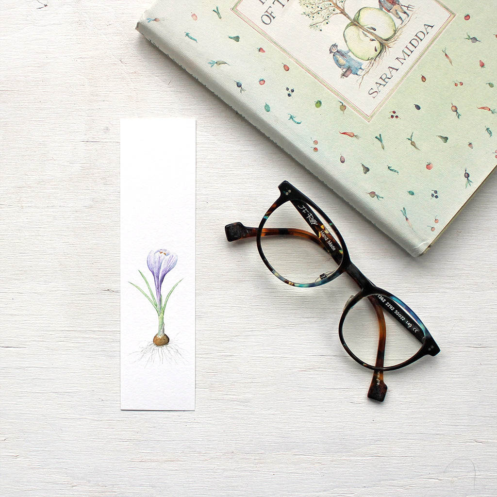 A paper bookmark featuring a watercolor painting of a striped purple crocus bulb. Artist Kathleen Maunder.