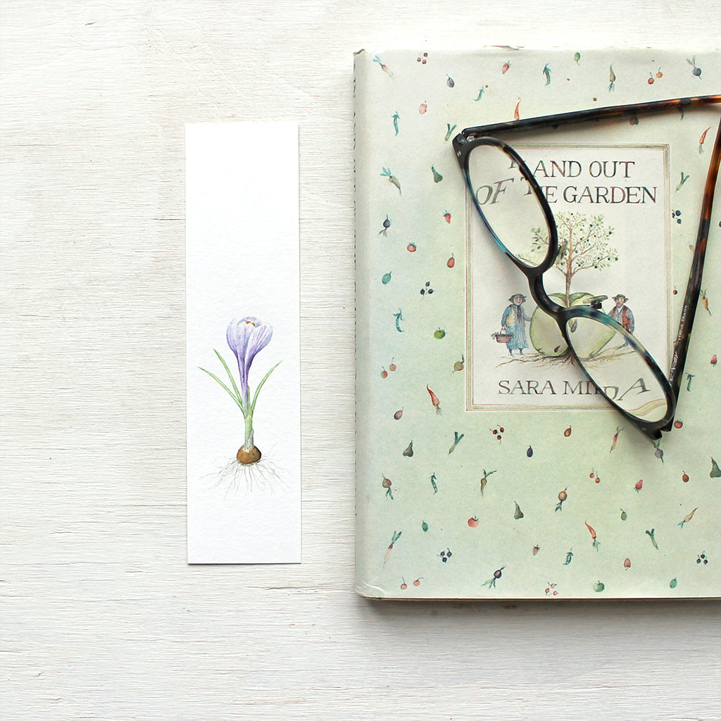 A paper bookmark featuring a watercolor painting of a striped purple crocus bulb. Artist Kathleen Maunder.