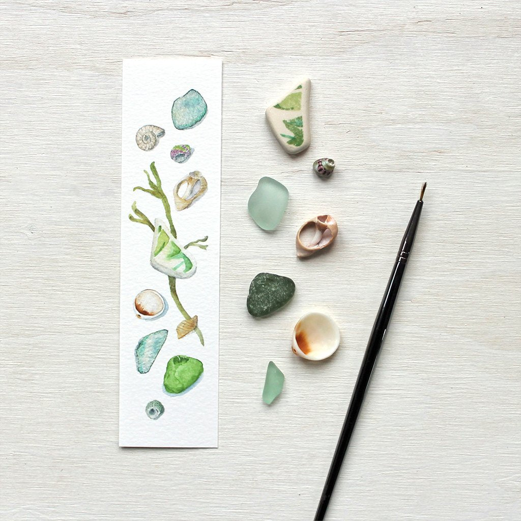 Paper bookmark with watercolour painting of beach finds: a collection of tiny seashells, sea glass, a worn ceramic fragment and a piece of seaweed.