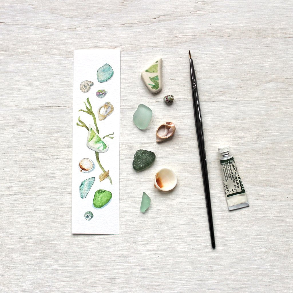 Paper bookmark with watercolor painting of a collection of sea finds including shells and beach glass. Artist Kathleen Maunder.