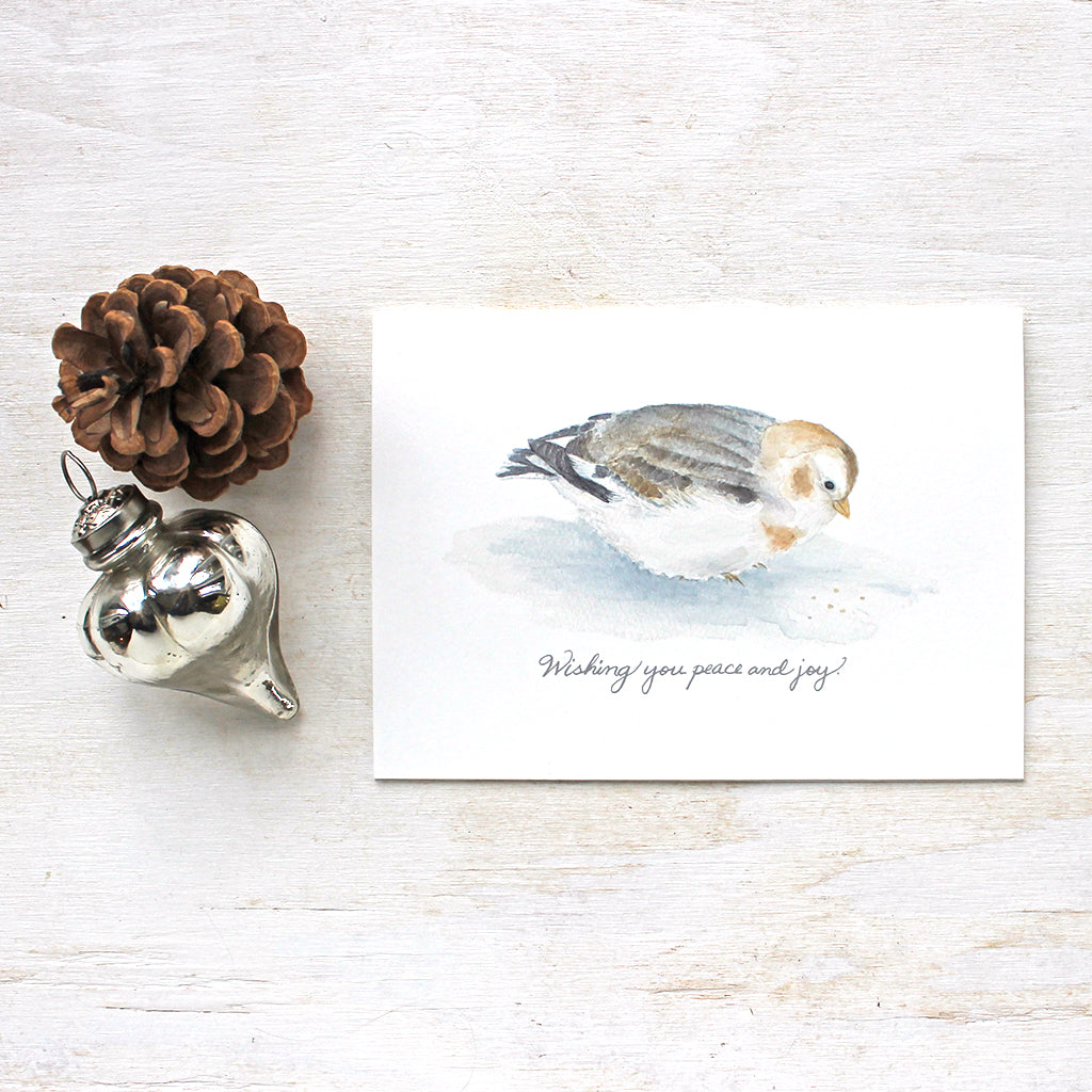A holiday card for bird lovers featuring a delicate watercolor painting of a snow bunting and the hand lettered greeting: 'Wishing you peace and joy'.