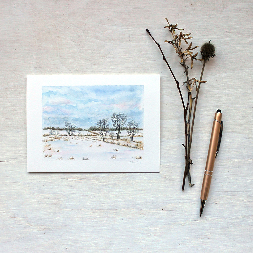 A note card featuring a watercolor painting of a winter landscape including snowy fields, trees, and a cloudy sky. Artist Kathleen Maunder.