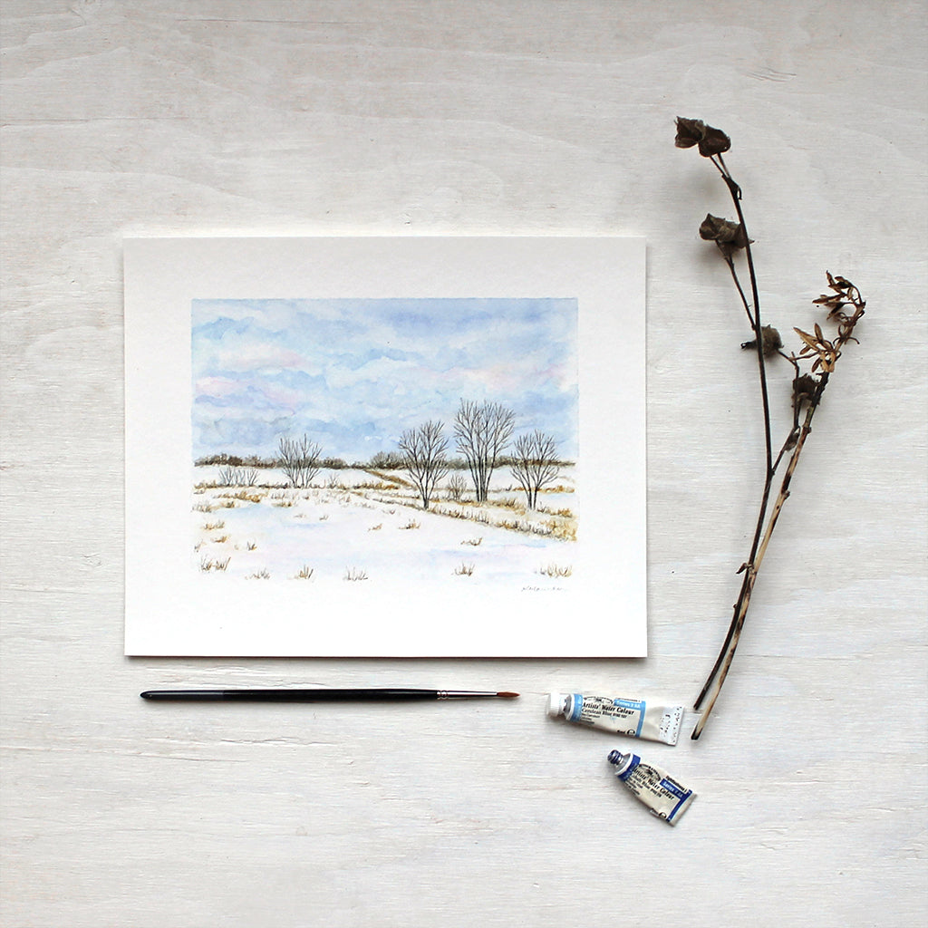 A print featuring a landscape painting in watercolor. Snowy fields, trees and blue wintry clouds. Artist Kathleen Maunder.