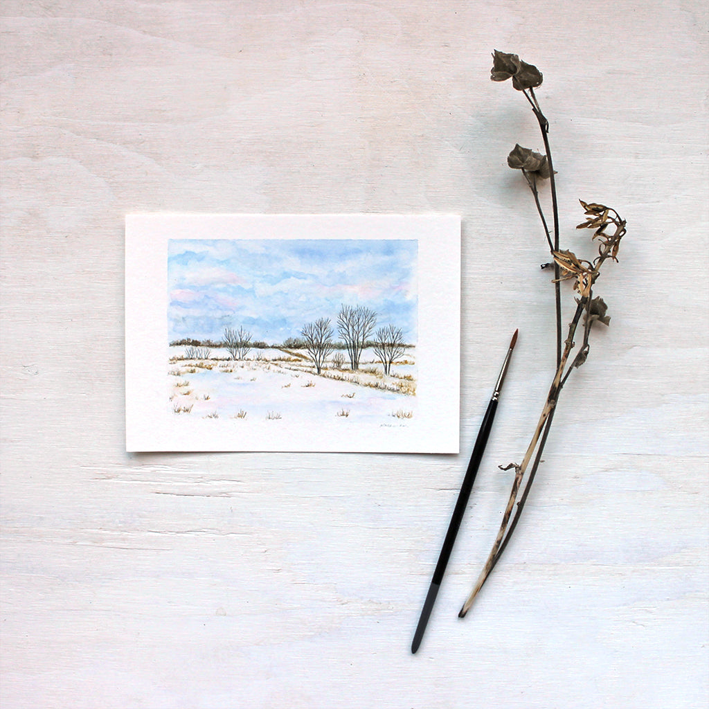 A print featuring a watercolor painting of a rural landscape: snowy fields, trees and blue wintry clouds. Artist Kathleen Maunder.