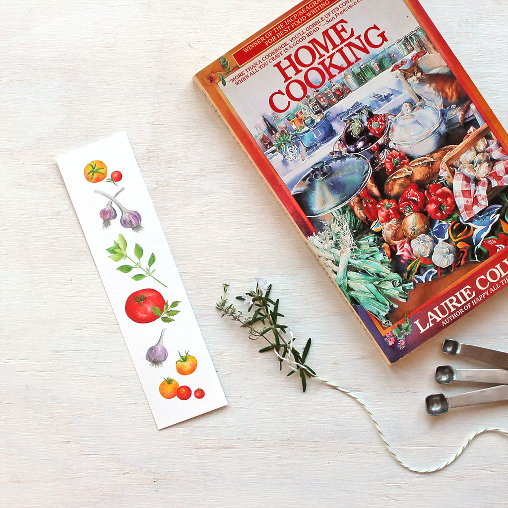 A bookmark featuring watercolor paintings of yellow and red garden tomatoes, purple-striped garlic and basil. Artist Kathleen Maunder.