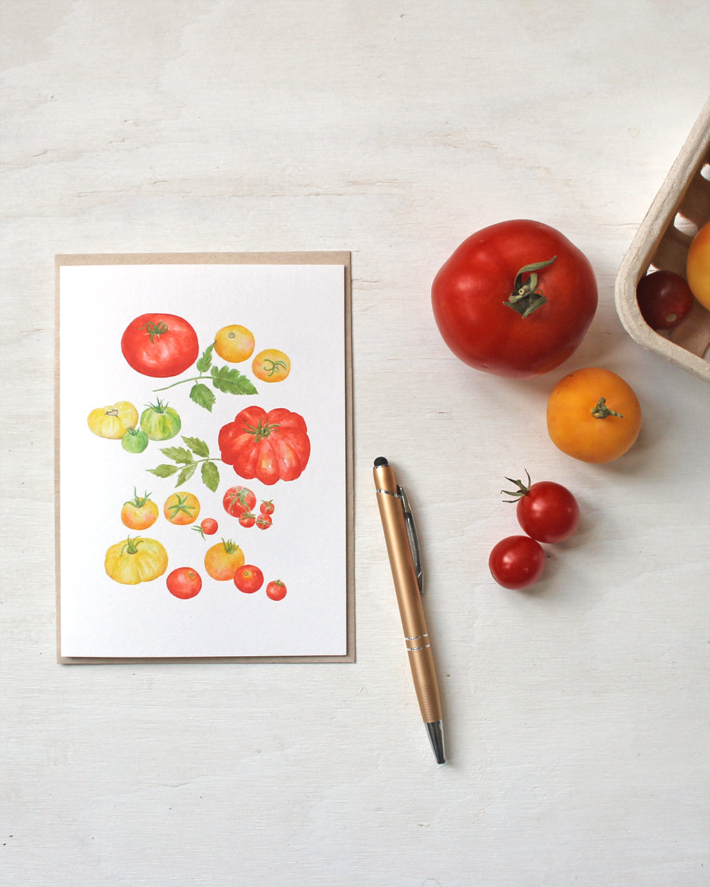 A blank note card featuring a watercolor painting of several types of red, yellow and green heirloom garden tomatoes. Artist Kathleen Maunder.