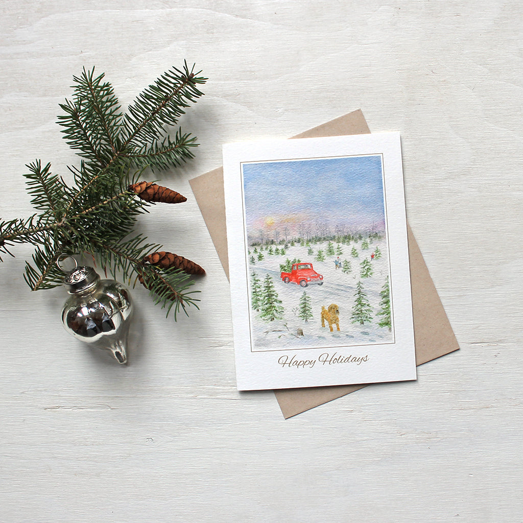 Holiday card featuring a watercolor of a Christmas tree farm with a red truck in the background and a dog in the foreground. Artist Kathleen Maunder