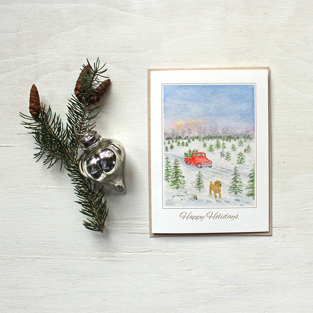 Holiday card featuring a watercolor of a Christmas tree farm with a red truck in the background and a dog in the foreground. Artist Kathleen Maunder