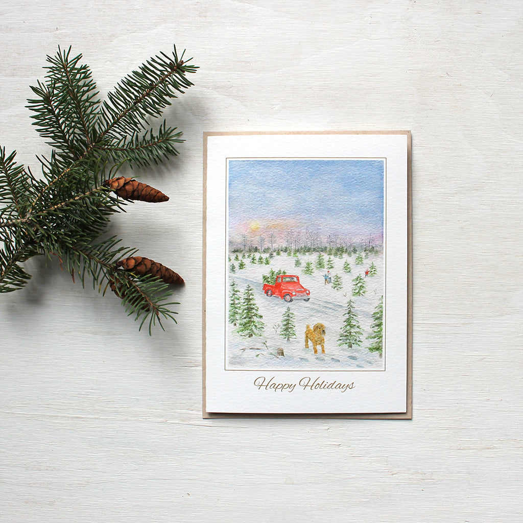 Holiday card featuring a watercolor of a Christmas tree farm with a red truck in the background and a dog in the foreground. Artist Kathleen Maunder