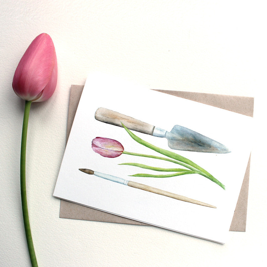 A note card featuring a watercolor painting of a garden trowel, a pink tulip and an artist's paintbrush. Artist Kathleen Maunder.