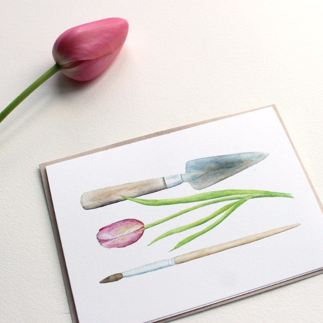 A note card featuring a watercolor painting of a garden trowel, a pink tulip and an artist's paintbrush. Artist Kathleen Maunder.