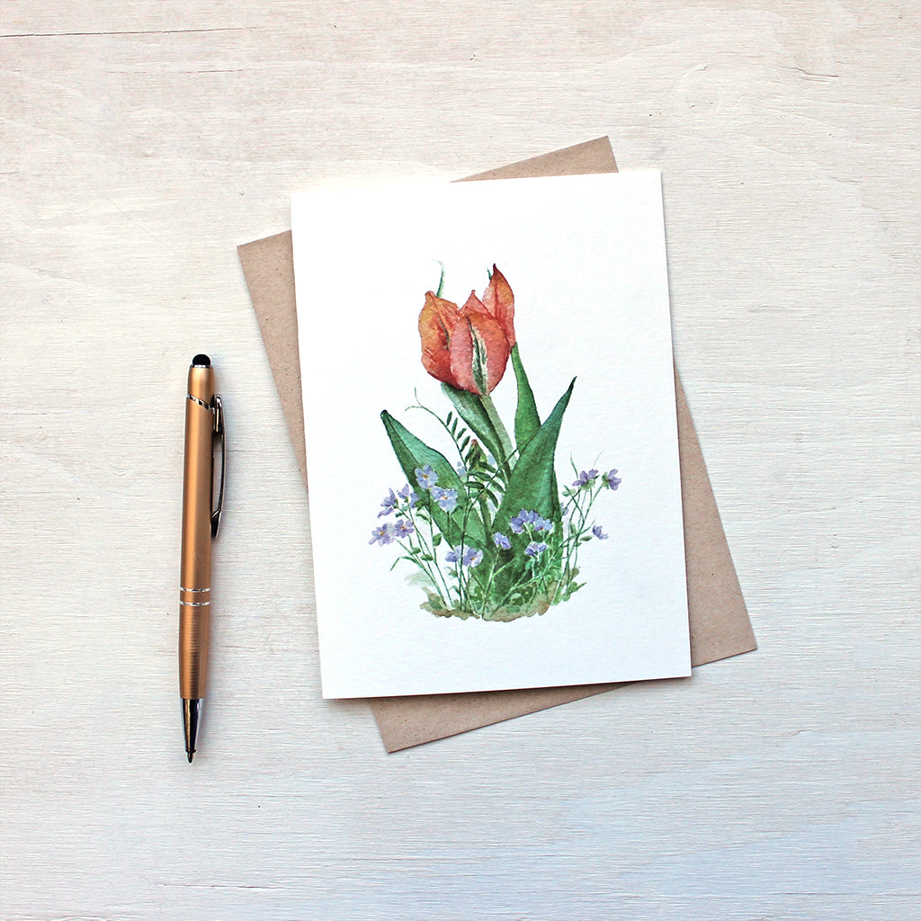 A lovely note card featuring a watercolor painting of an orange-red tulip surrounded by violets. Artist Kathleen Maunder.