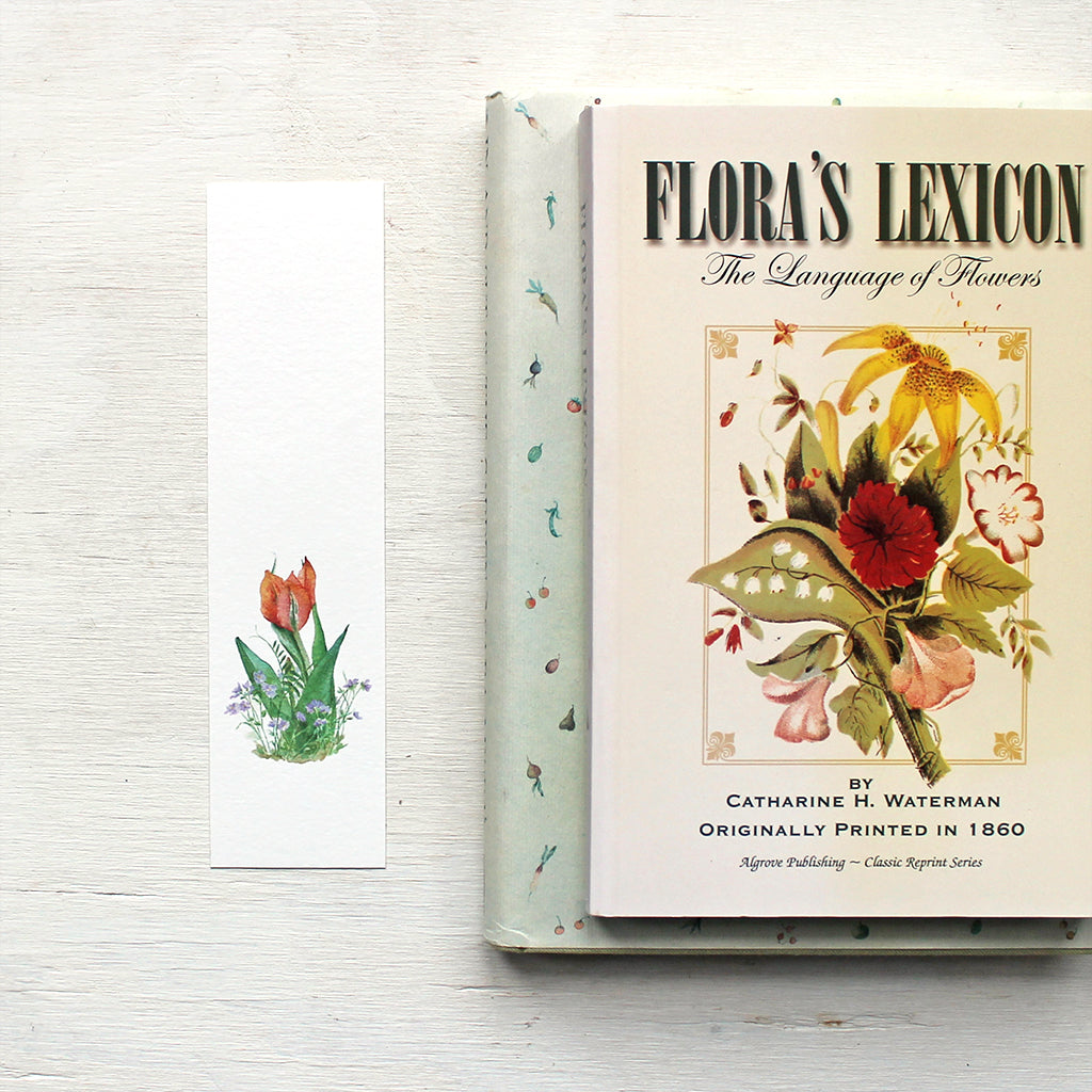 A paper bookmark featuring a watercolour painting of an orange red tulip surrounded by violets. Artist Kathleen Maunder.