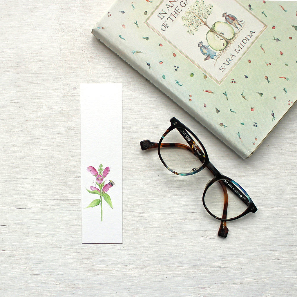 A floral bookmark featuring a watercolor painting of pink turtlehead (chelone) and a bee. Artist Kathleen Maunder.