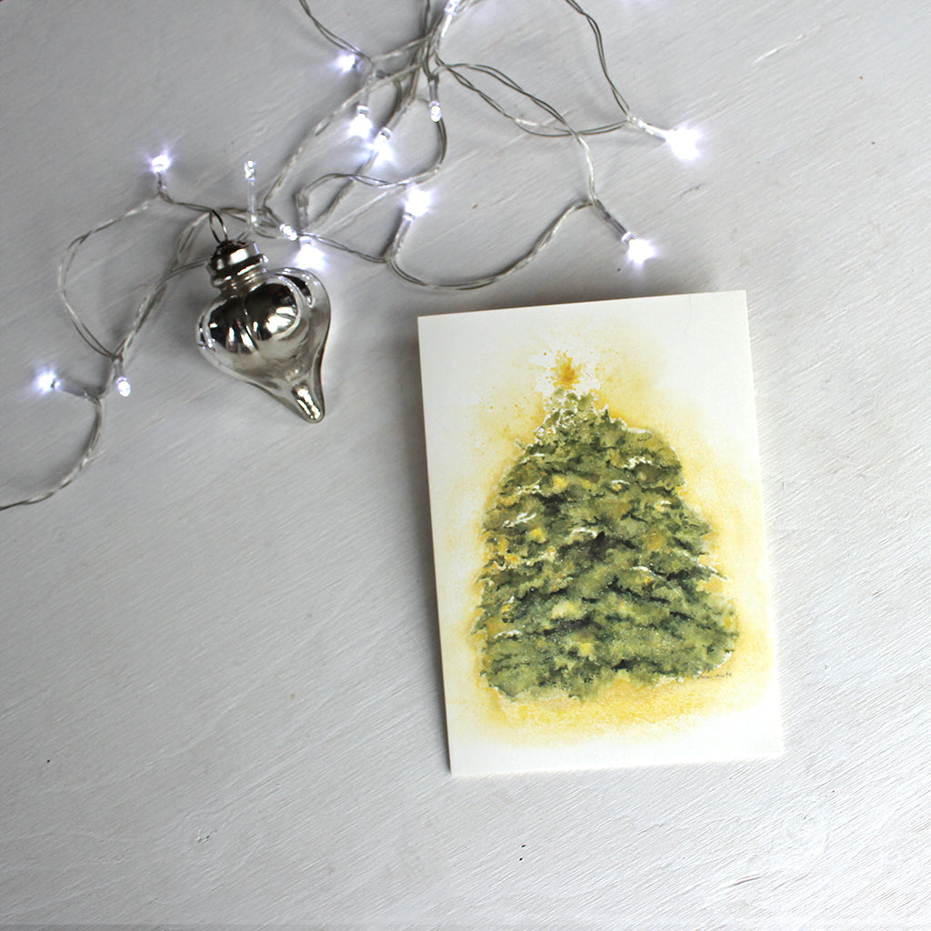 A holiday card featuring a watercolor of an evergreen tree dusted with snow, topped with a star, and covered in golden light. Artist Kathleen Maunder