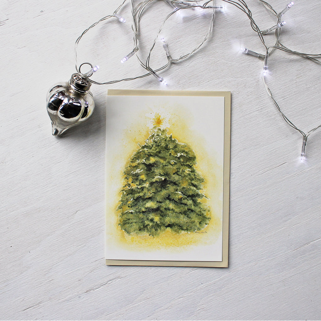 A holiday card featuring a watercolor of an evergreen tree dusted with snow, topped with a star, and covered in golden light. Artist Kathleen Maunder