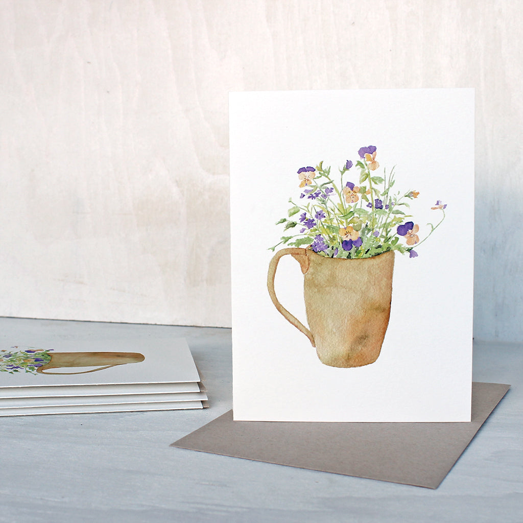 A note card featuring a watercolor painting of a bouquet of yellow and purple violas and purple verbena in a ceramic mug. Artist Kathleen Maunder