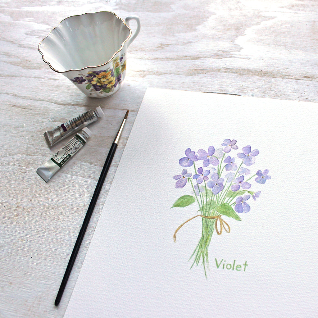 Art print featuring a delicate watercolor painting of a small bouquet of violets tied with a string. Artist Kathleen Maunder. (A tea cup, paint and paintbrush in background.)
