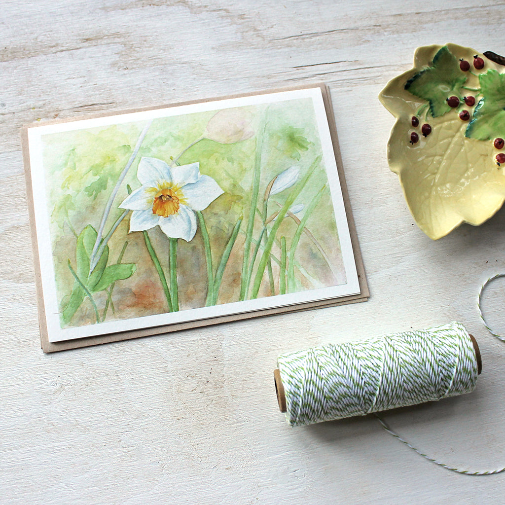 A beautiful note card featuring a watercolor painting of a single white and yellow daffodil (narcissus) in a spring garden. Artist Kathleen Maunder.