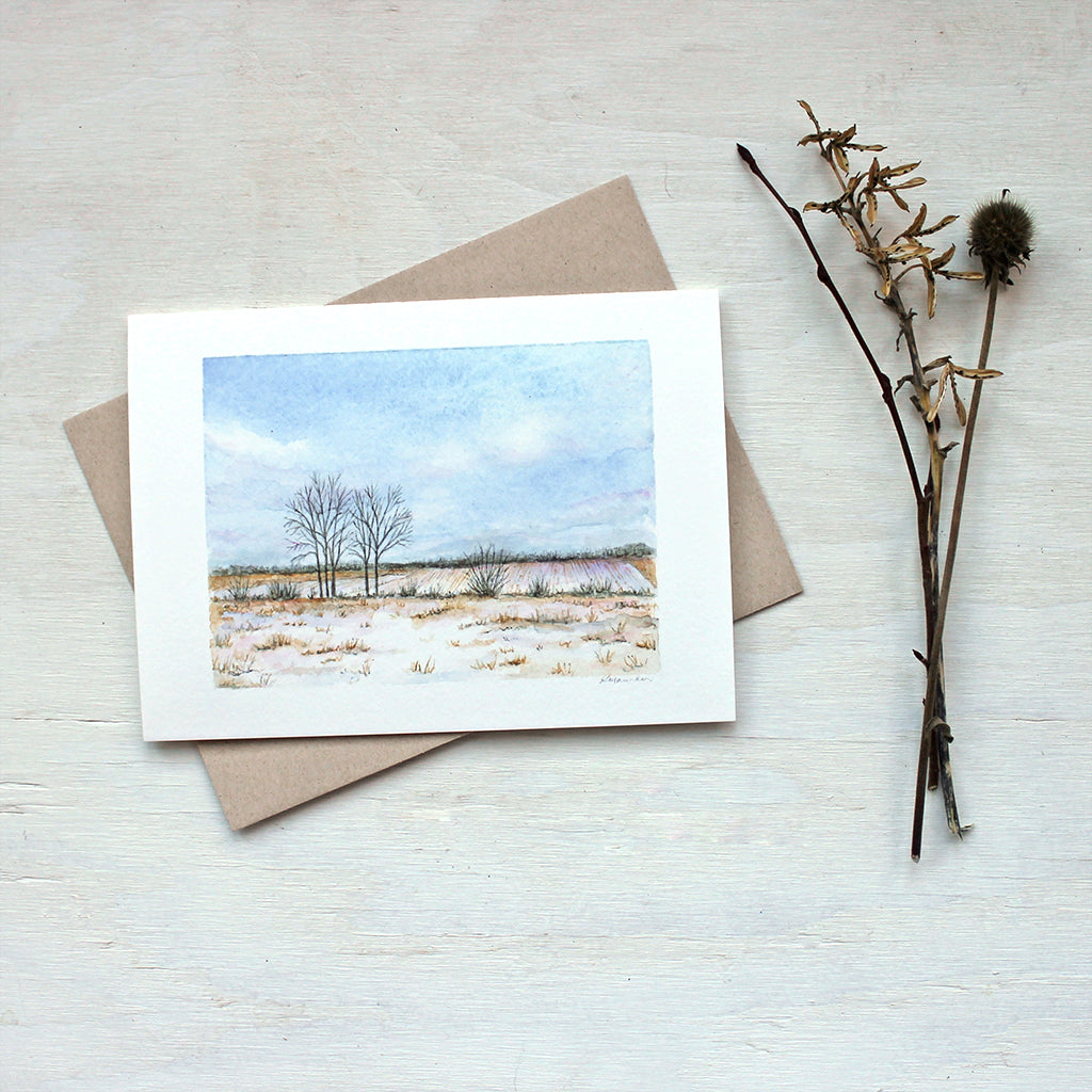 A note card featuring a watercolor painting of a winter landscape including a snowy field, trees, blue sky and fluffy clouds.