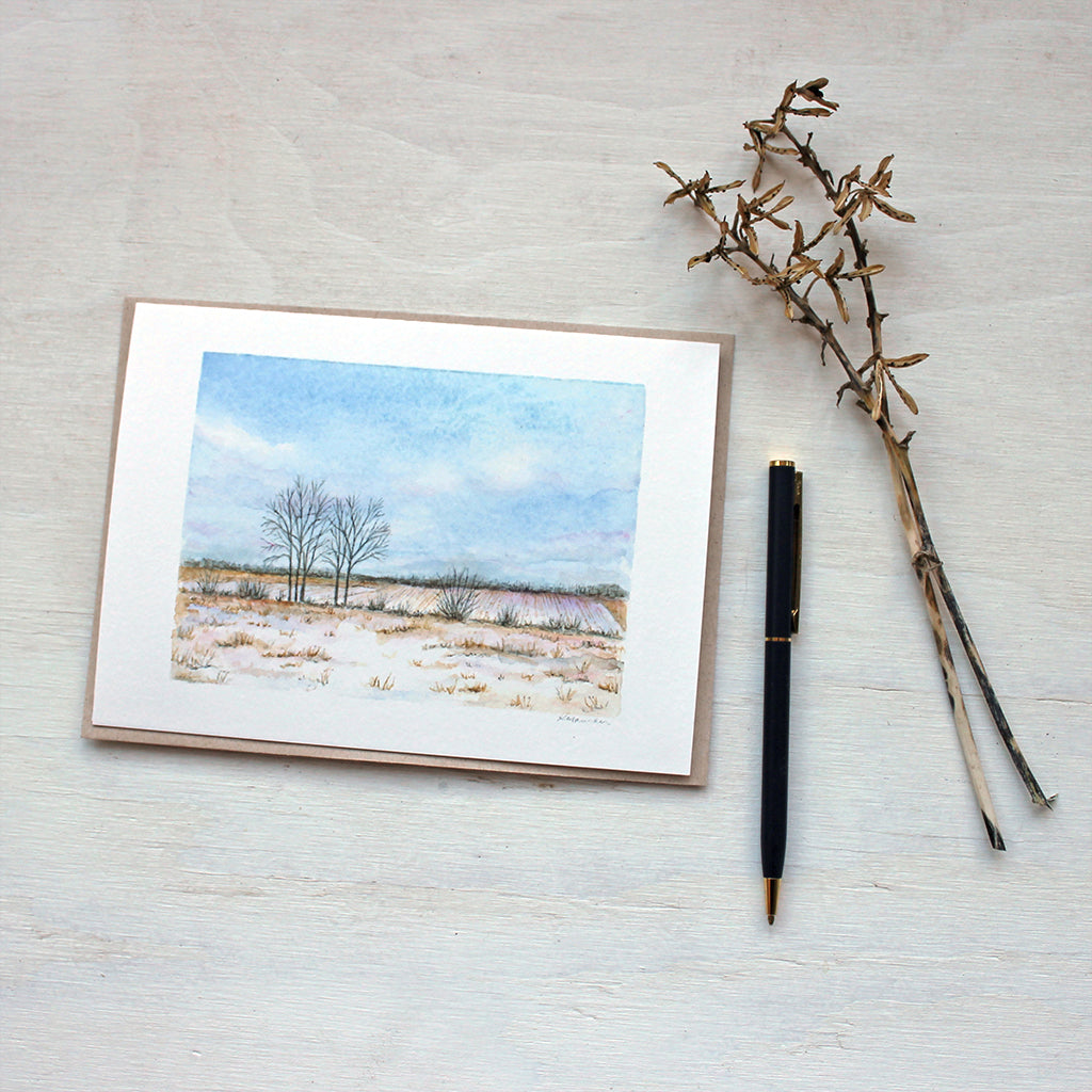 A blank note card featuring a watercolor painting of a snowy rural landscape. Artist Kathleen Maunder.
