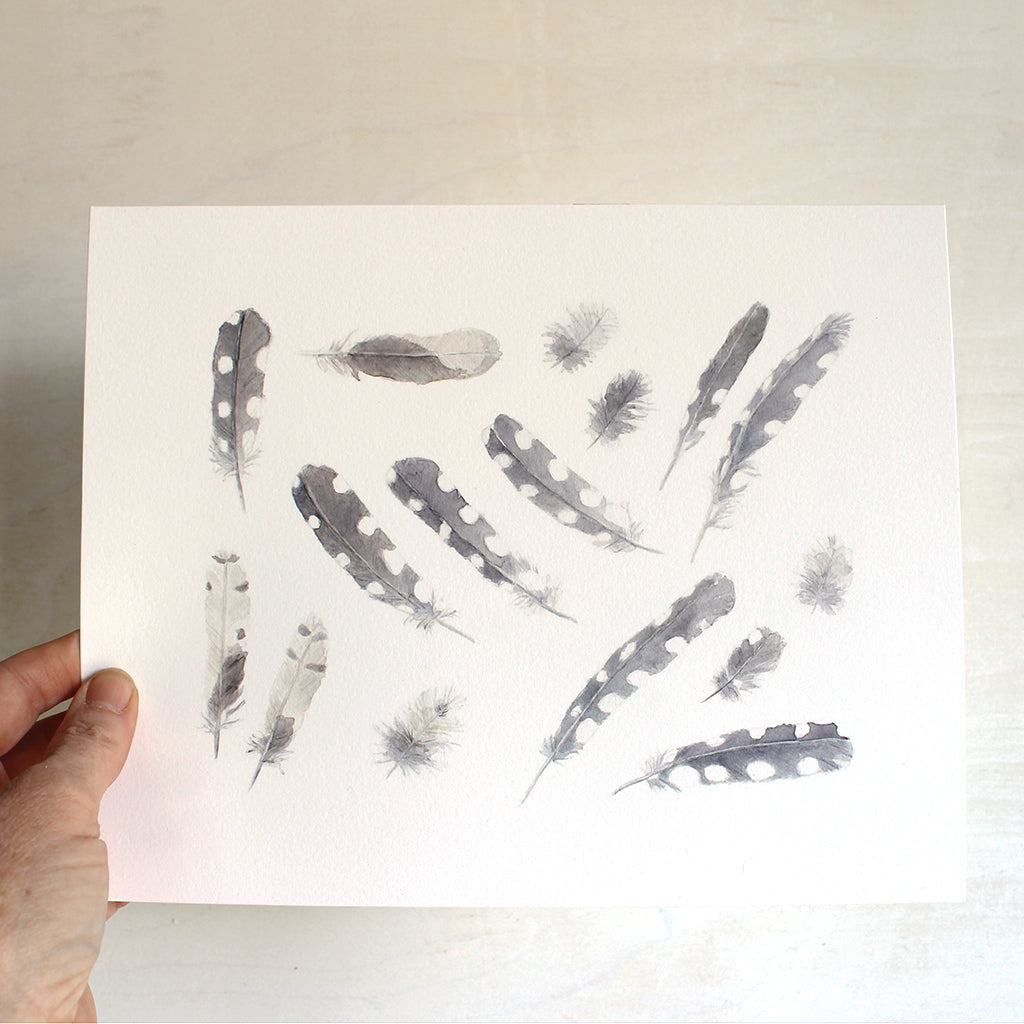 Artist holding an art print featuring a delicate watercolor painting of sixteen woodpecker feathers. 
